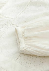 Tissu blanc brodé avec un motif texturé, doté de manches froncées et de détails découpés complexes le long des coutures.