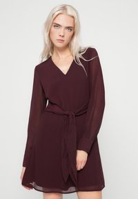 Μπρούμυτα φόρεμα μακρυμάνικο με V-λαιμόκοψη και ζώνη στη μέση. Κατασκευασμένο από ελαφρύ ύφασμα, διαθέτει φούστα με ροή και κουμπωτές μανσέτες.