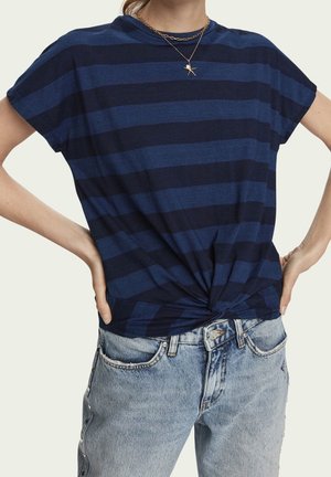 Woman wearing navy and black striped short-sleeve top knotted at waist, light blue jeans with side studs, and layered gold necklaces.