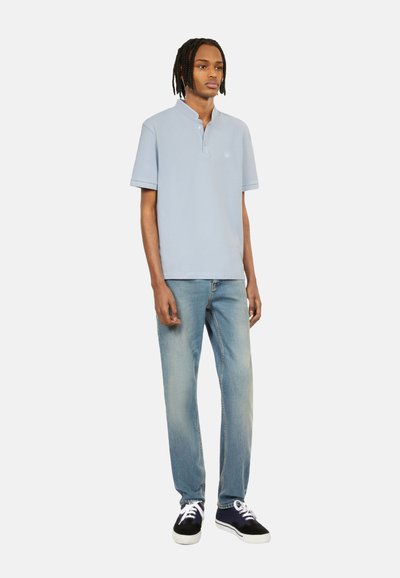 Polo shirt azzurro chiaro con colletto discreto, abbinato a jeans blu sbiaditi e sneaker scure. Tessuto morbido con dettaglio del logo sul petto.