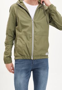 Olivgrüne Zip-Jacke mit Kapuze, elastischen Bündchen und Saum. Das Material wirkt leicht und hat eine glatte Textur. Weißer Reißverschluss und auffällige Etiketten sind sichtbar.