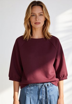 Frau mit schulterlangem blondem Haar, die ein burgunderfarbenes Dreiviertelarm-Top und hoch taillierte blaue Jeans mit gebundenem Gürtel trägt.