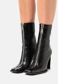 Bottes en cuir noires mi-mollet avec fermetures éclair argentées à l'avant et talons blocs, portées sur des jambes nues sur un fond blanc.