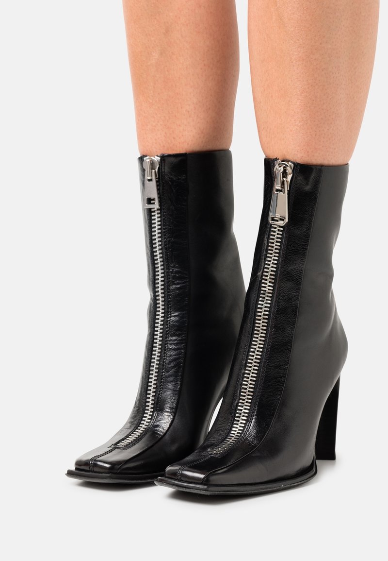 Bottes en cuir noires mi-mollet avec fermetures éclair argentées à l'avant et talons blocs, portées sur des jambes nues sur un fond blanc.