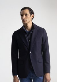 Blazer azul marino con tejido texturizado, dos bolsillos frontales y cierre de un solo botón, superpuesto sobre un chaleco oscuro y una camisa azul.