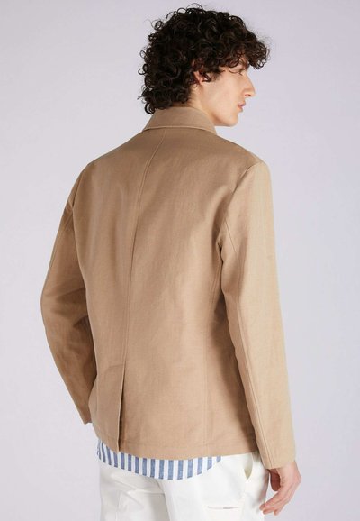 Beige Jacke mit einer strukturierten Design und subtiler Textur. Verfügt über einen Kragen und einen Rückenbelüftung, getragen über einem gestreiften Hemd und hellen Hosen.