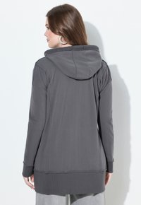 Woman with brown hair wearing large hoop earrings and a long dark gray hooded sweatshirt, shown from the back.