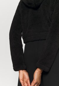 Svart croppad hoodie i ett texturerat, fluffigt material. Har en huva och långa ärmar med åtsittande mudd, bärs över en svart outfit.