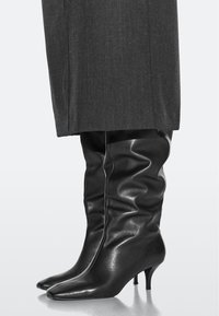 Stivali neri al ginocchio in pelle lucida, caratterizzati da un design morbido, punta appuntita e tacco alto e sottile.