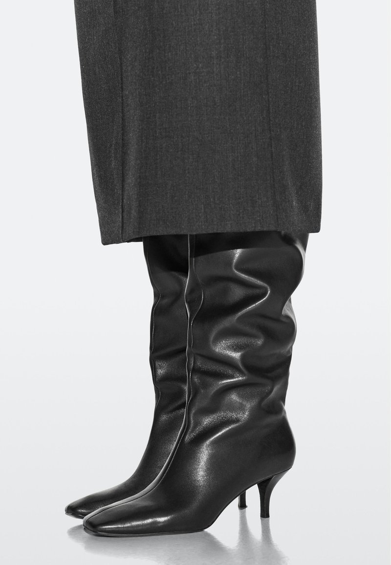 Stivali neri al ginocchio in pelle lucida, caratterizzati da un design morbido, punta appuntita e tacco alto e sottile.