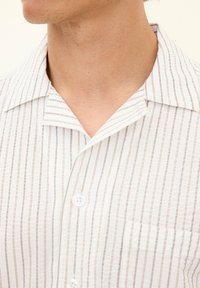 Biała koszula krótko-sleeve z cienkimi, pionowymi, brązowymi paskami, z kołnierzykiem typu camp, guzikami z przodu i kieszonką na lewej piersi. Teksturowany materiał.