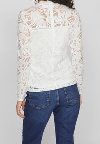 Top bianco in pizzo a maniche lunghe con motivi floreali, colletto alto e chiusura a bottone sul retro, abbinato a jeans in denim blu.