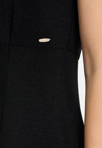 Schwarzes Kleid aus einem strukturierten Stoff mit einem leichten Schimmer. Ein kleines, metallic glänzendes Markenetikett befindet sich in der Nähe des Ausschnitts.