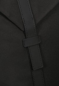 Musta kangaskassi, jossa on sileä nahkaremmi ja solki. Raskaampi pinta ja puhtaat linjat määrittävät sen modernin, minimalistisen muotoilun.