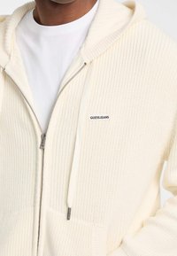 Kremasta pletena dukserica s kapuljačom, s rebrastom teksturom, zatvaranjem na patentni zatvarač i prednjim džepovima. Ima malu oznaku "GUESS JEANS" s lijeve strane.