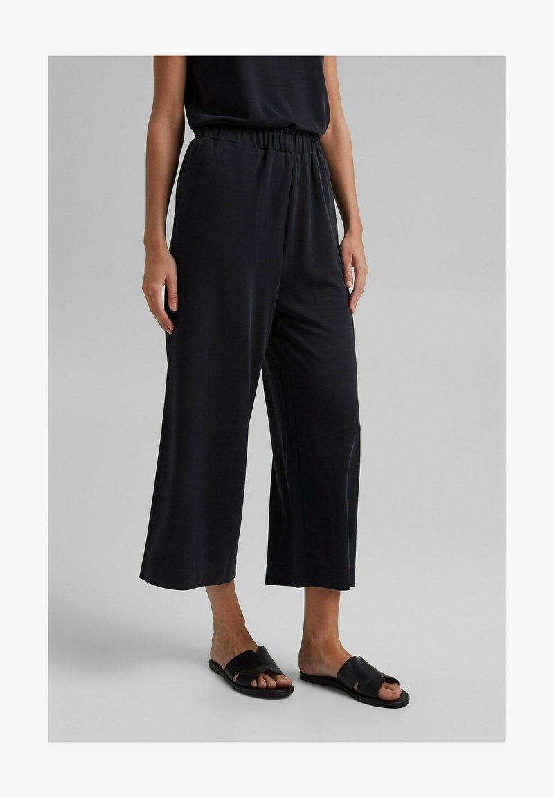 Pantalon noir à jambes larges en tissu lisse, doté d'une taille élastique et d'une coupe décontractée, porté avec des sandales plates noires.