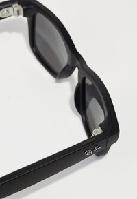Schwarze Plastiksonnenbrille mit dunklen Gläsern, quadratischer Form, silbernen Hardware-Akzenten und einem weißen Ray-Ban-Logo an der Schläfe.