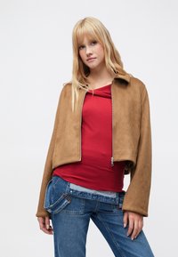 Brun jakke i imiteret ruskind med kort design, stor krave og lynlås, kombineret med en rød top og blå denimjeans.