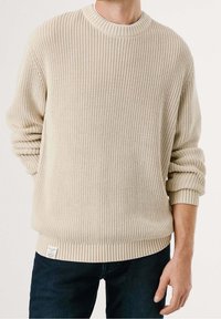 Pull beige tricoté avec une texture côtelée, col rond et poignets retroussés, associé à un jean denim foncé. Étiquette visible à l'ourlet.