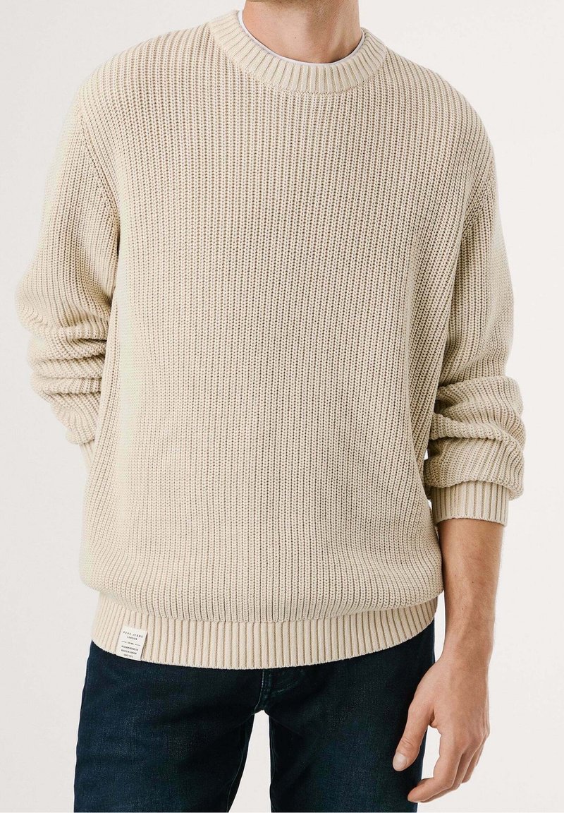 Pull beige tricoté avec une texture côtelée, col rond et poignets retroussés, associé à un jean denim foncé. Étiquette visible à l'ourlet.