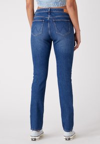 Osoba oblečená v modrých džínsech a světle modrých teniskách, stojící zády k fotoaparátu na neutrálním pozadí.