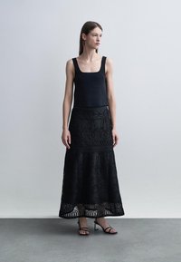 Femme portant un haut noir sans manches à col carré, une longue jupe noire à motif en dentelle, et des talons noirs à brides, debout contre un mur gris clair.