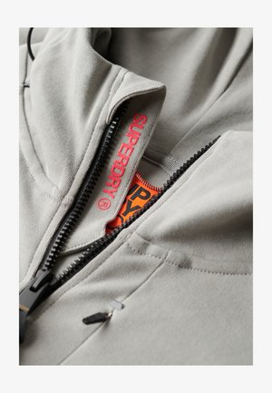 Sudadera gris con cremallera que presenta una textura suave, cremallera negra y detalle del logo "SUPERDRY" en rojo en la solapa interior.
