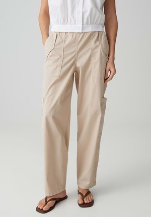 Beige Weite-Hosen mit Front- und Seitentaschen, getragen mit einem weißen ärmellosen Top und braunen Sandalen, stehend vor einem schlichten Hintergrund.