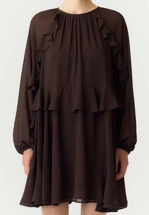 Femme portant une robe longue à manches longues brun foncé avec des détails à volants sur la poitrine, les manches et l'ourlet, debout devant un fond uni.