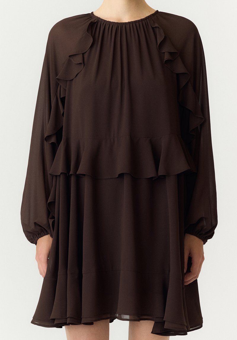 Femme portant une robe longue à manches longues brun foncé avec des détails à volants sur la poitrine, les manches et l'ourlet, debout devant un fond uni.
