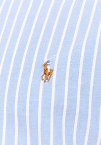 Jasnoniebieska koszula w paski z małym, brązowym i białym haftowanym logo konia z jeźdźcem. Gładka tekstura, klasyczny krój kołnierza.