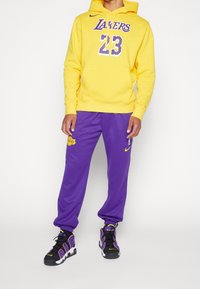 Hoodie jaune avec graphique "Lakers" et "23". Pantalons de survêtement violets avec logo. Baskets noires et violettes avec semelle blanche.