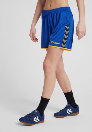 Person iført blå sports shorts med gult kantbånd og sorte chevronstriber, kombineret med sorte sokker og blå sneakers med hvide chevroner.