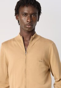 Jonge man met korte dreads, gekleed in een beige zip-up trui met een klein "BOSS" logo op de borst, recht tegemoet kijkend tegen een effen achtergrond.