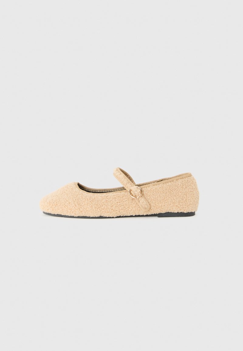 Beige luddig Mary Jane plat sko med rund tå, justerbar rem med cirkulär spänne och svart sula på vit bakgrund.