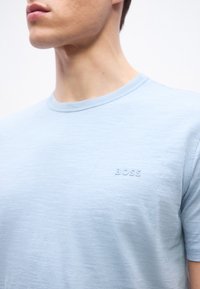 Άνδρας που φοράει ανοιχτό μπλε μπλουζάκι με στρογγυλή λαιμόκοψη και μικρό ανάγλυφο λογότυπο "BOSS" στο αριστερό μέρος του στήθους, στέκεται μπροστά σε λευκό φόντο.