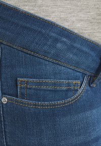 Vaqueros de mezclilla azul oscuro con un pequeño bolsillo trasero, costuras amarillas y un botón plateado. La textura de la tela es visible con un ligero desgaste.