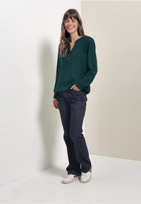 Blusa verde con escote en V y mangas largas, combinada con jeans oscuros y zapatillas blancas. Textura de tela suave y corte relajado.