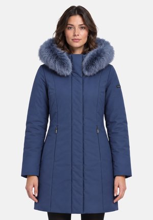 Donna con un lungo cappotto invernale blu con un grande cappuccio soffice bordato di pelliccia, in piedi su uno sfondo semplice.