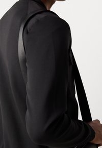 Chemise noire à manches longues avec une texture lisse et une coupe ajustée, avec une bandoulière d'un sac noir reposant sur le tissu.