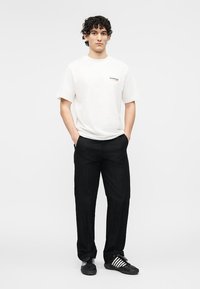 Junger Mann steht mit Händen in den Taschen, trägt ein weißes kurzärmeliges Hemd, schwarze Hose und schwarze Turnschuhe mit weißen Streifen.