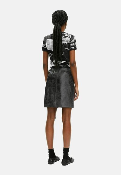 Vrouw met lang gevlochten haar, gekleed in een zwart-wit tijdschriftprint top, zwarte leren rok, zwarte sokken en gestifte zwarte schoenen, staand.