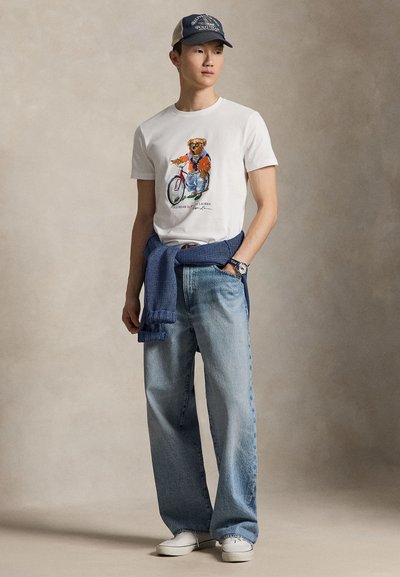 Ung mann iført hvit t-skjorte med bjørnegrafikk, lyseblå jeans, hvite joggesko, blå genser knyttet rundt livet og marineblå baseballcaps.