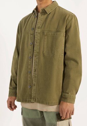 Mężczyzna w oliwkowo-zielonej kurtce na guziki z kieszenią na piersi oraz spodniach cargo z naszywkami, stojący na tle jasnego, jednolitego tła.