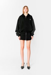 Sudadera negra de gran tamaño con cremallera frontal, con un tirador circular y un logo bordado. Combinada con una falda corta negra y tacones.