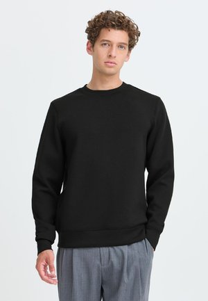 Jonge man met krullend haar die een zwarte trui met ronde hals en lange mouwen draagt en grijze geplooide broek, staande tegen een effen achtergrond.