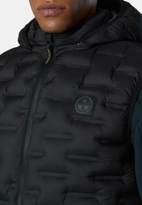 Chaleco de plumas negro con textura acolchada, cuello alto y cremallera frontal. Tiene un parche de logotipo redondo en la parte izquierda del pecho.