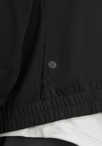 Chaqueta ligera negra con dobladillo fruncido, fabricada en material suave, con un detalle sutil del logo cerca de la costura. Lleva una capa sobre una camiseta blanca.