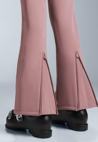 Ružičaste hlače trapeznog kroja od glatkog tkanja s bočnim patentnim zatvaračima na rubu. Uparene s masivnom crnom obućom s metalik detaljima.