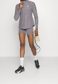 Langarmiges graues Sportoberteil mit halbem Reißverschluss, kombiniert mit passenden grauen Shorts; trägt schwarze und weiße Sneakers und hält ein Springseil in der Hand.